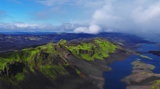 Aerial shot sweeps over Iceland Highlands near Landmannalaugar, gliding along ridges and lake corridor with lava fields, craters, clouds, and a faint rainbow.