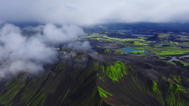 Aerial drone sweep over Iceland volcanic highlands shows black lava, green moss, sharp ridges, turquoise crater lakes, braided rivers, and low clouds in soft daylight.
