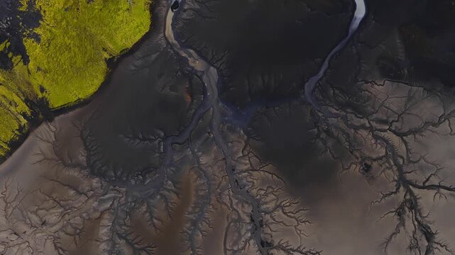 Top down aerial view shows braided milky blue meltwater over charcoal black sand and ash, with yellow green moss lava fields in Iceland, high contrast texture.