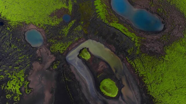 Aerial top down of south Iceland volcanic terrain, neon green moss on black lava, eye shaped crater ponds, vivid blue lakes, braided meltwater, sinuous turquoise river.