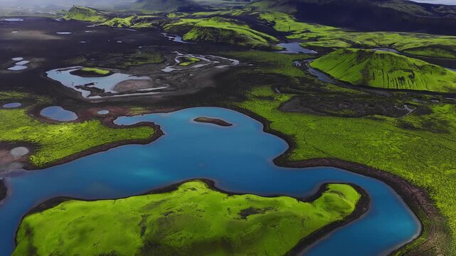 Aerial sweep over Iceland volcanic highlands shows turquoise lakes, winding rivers, mossy craters, tuff hills, and black lava fields in summer with soft horizon mist.