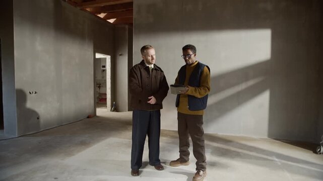Full length shot of Black male interior designer holding material samples, discussing design ideas and layouts with client at unfinished construction site for renovation or interior project
