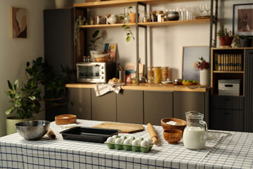 Kitchen table holding eggs, milk, flour, rolling pin, baking tray, mixing bowls, kitchen in background featuring shelves with plants, jars, books, and small appliances