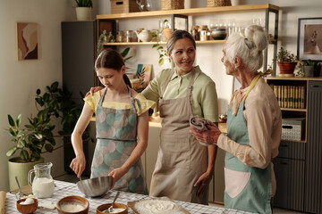 Caucasian senior woman talking with Caucasian middle aged woman while Caucasian teenage girl mixing ingredients in metal bowl on kitchen counter, all wearing aprons, surrounded by plants