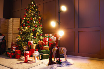 Decorated Christmas tree with many gifts, reindeer and glowing lamp near brown wall in living room at night