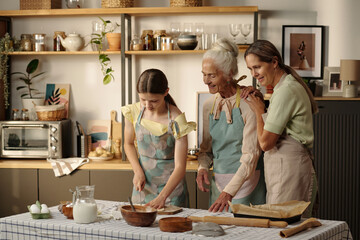Caucasian teenage girl preparing dough while middle aged Caucasian woman and senior Caucasian woman standing beside her smiling in modern kitchen, engaging in baking activity together