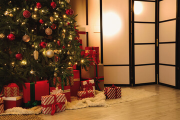 Decorated Christmas tree with many gifts, folding screen and glowing lamps in living room at night