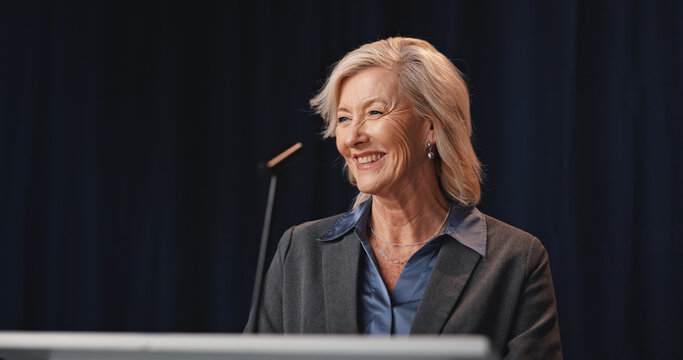 Politics, smile and speech with mature woman on stage for democracy, election or government campaign. Happy, microphone and podium with speaker at conference, meeting or summit for support or vote