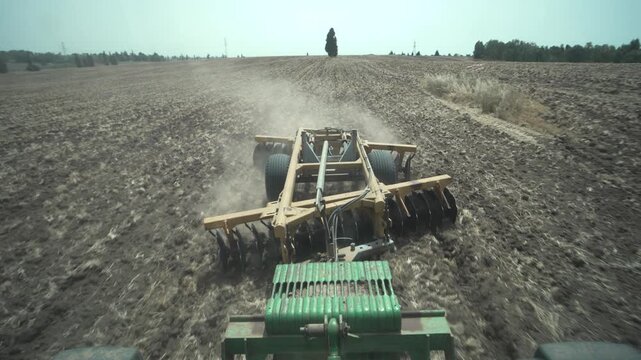 A disc harrow plows a dry field under bright daylight, raising dust clouds. Blue summer skies. Field ready for sowing. Shot from tractor driver POV. Modern agriculture in Israel, the Holy land