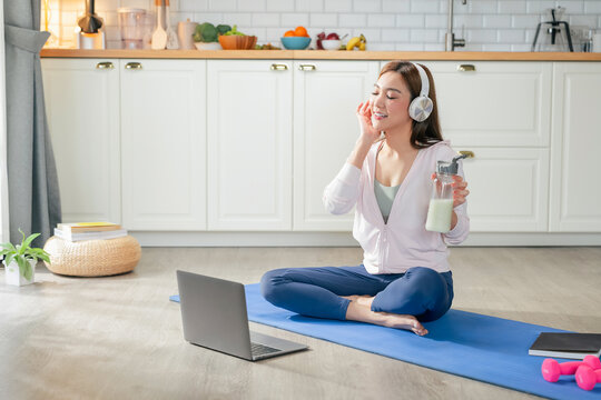 happy motivated Asian woman practices yoga at home online private class stretching body perfect balance listening instructor headphones holds protein shake embracing healthy lifestyle peaceful home