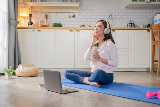 happy motivated Asian woman practices yoga at home online private class stretching body perfect balance listening instructor headphones holds protein shake embracing healthy lifestyle peaceful home