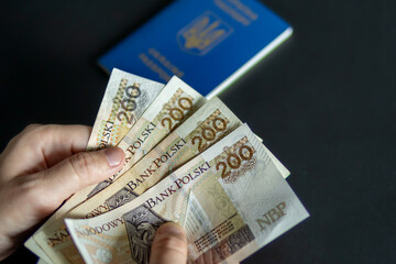 A hand holding several 200 PLN Polish banknotes with a Ukrainian passport in the background. Symbol of money, migration, border crossing