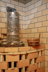 Fototapeta premium Vertical shot of a brick-covered sauna stove with hot stones, with a water ladle placed on the edge. A warm brick wall in the background creates a rustic and traditional steam room atmosphere.