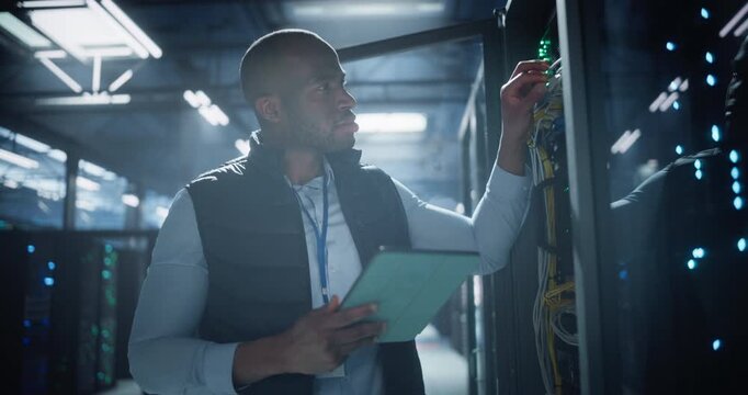 Data center worker uses a tablet to monitor server rack performance, checks network cables, and inspects illuminated hardware in facility filled with servers. Portrait looking at camera