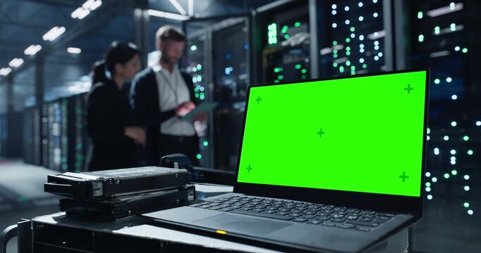 Laptop with green screen chroma key sits on a cart beside hard drives in data center.  Two people discuss network infrastructure and server maintenance among  server racks and mainframes.