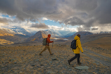 Two hikers with backpacks and trekking poles walk a mountain trail during the golden hour....