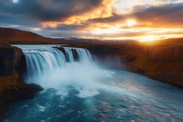 majestic waterfall flowing into a wide river under a dramatic sunset sky with clouds and distant snowy mountains