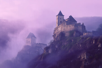Medieval Castle Hohenwerfen sits high on a cliff, partially obscured by heavy fog at sunrise in Werfen, Austria. The stone walls of the castle stand out against the pink sky.