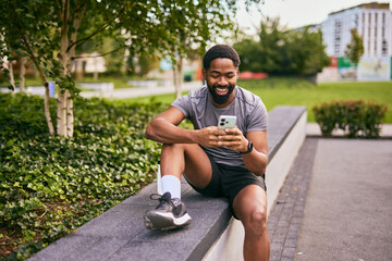 Obraz premium Happy Man in Athletic Wear Texting on Phone While Sitting on Park Edge