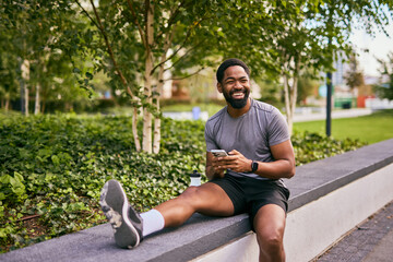 Obraz premium Smiling Man in Workout Gear Using Phone Outdoors While Relaxing on Park Ledge