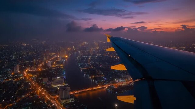 Time-lapse view from airplane window flying over Bangkok at night. The Chao Phraya River glows with reflections from city lights as cars create golden trails along the illuminated streets below.