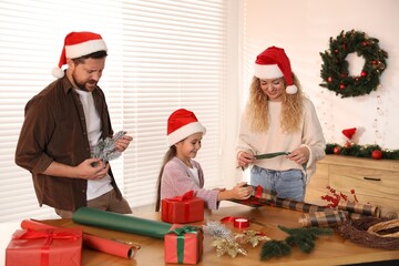Happy family in Santa hats wrapping Christmas gifts at table indoors