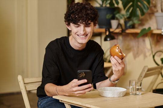 Young cheerful person enjoying a snack while smiling at their smartphone in a cozy cafe