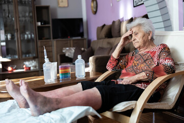 An elderly woman sitting in a chair with a tired expression, surrounded by medical supplies, conveying a sense of fatigue and health challenges in a warm, domestic environment.