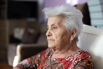 An elderly woman sits quietly in her living room, lost in thought, reflecting on memories and experiences while gazing into the distance with a calm demeanor and expression.