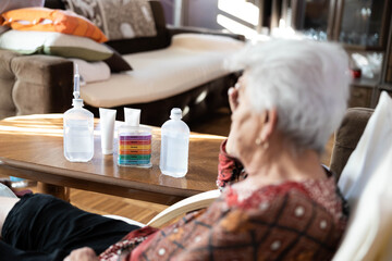 An elderly woman sits in a chair, observing bottles of skincare products on a table while exuding a sense of peace, illustrating the connection between self-care and well-being in later years.