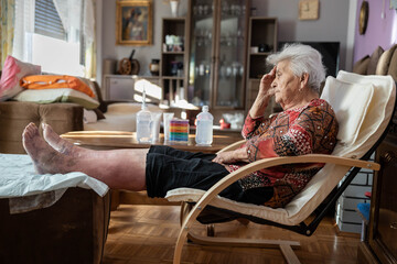 The elderly woman sits on her chair, with her legs raised and medical supplies placed strategically on the table beside her, conveying a sense of care and attention to her health.