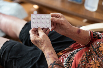 An elderly woman with visible hands showing a pack of medication, highlighting self-care and medication management in a warm and comforting home environment filled with personal items.