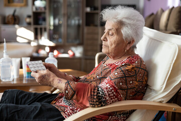 A senior woman holding a pack of pills in her hands, sitting thoughtfully in her armchair, reflecting on her health journey, emphasizing the value of self-care and awareness in her life.