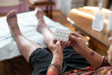 An elderly woman thoughtfully examining a pack of pills in her lap while resting in a cozy chair, representing careful medication management and introspection in a family-oriented setting.