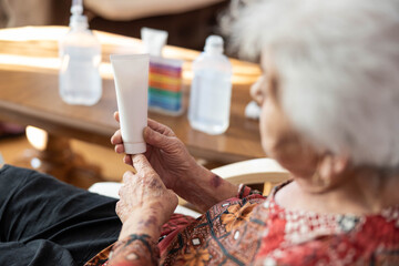 An elderly woman examines a tube of cream, visibly focused on its contents. The setting suggests a moment of self-care and attention to skincare, highlighting age-related concerns.
