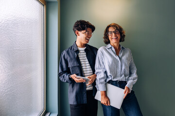 Two people laughing near a window, holding different electronic devices