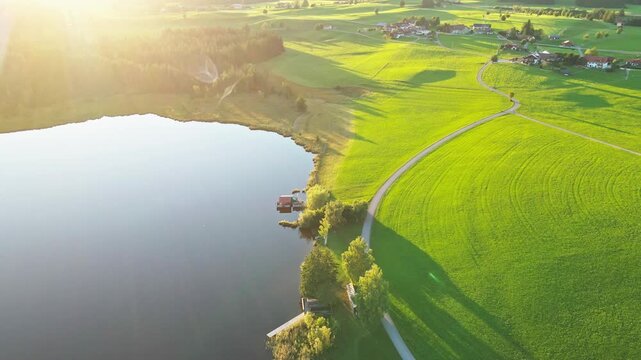 Aerial view of a serene lake mirroring the sky, bordered by vibrant green fields and a winding path under the golden sunlight, Nesselwang, Bayern, Germany.