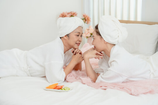 Senior Asian mother lying face to face with young daughter during skincare time on bed in relaxing home spa session both smiling warmly wearing towel wraps and white bathrobes indoor