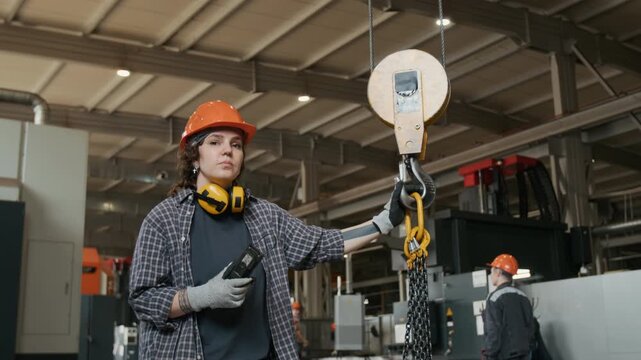 Portrait of young female shop floor supervisor in orange hard hat holding walkie talkie and hook with steel chain while coordinating lifts of heavy structures at metal fabrication plant, slow motion