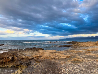 Calm evening by the rocky shore as waves gently crash under an expansive sky filled with dramatic clouds near the coastline