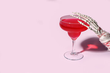 Skeleton hand with glass of tasty cocktail for Halloween celebration on pink background