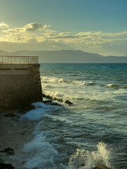 Fototapeta premium Waves crashing against a stone pier at sunset near a coastal town with mountains in the backdrop