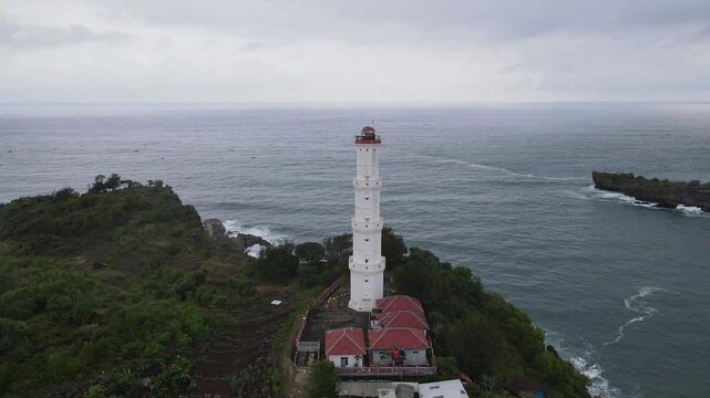 Aerial view of the Baron Lighthouse, a white beacon standing tall against the ocean's gray waves, surrounded by lush green cliffs, Yogyakarta, Yogyakarta, Indonesia.