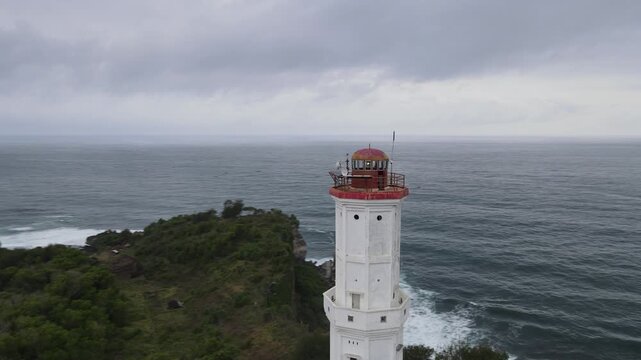 Aerial view of the white lighthouse at Baron Beach contrasts with the dark ocean and lush green coastline, Yogyakarta, Yogyakarta, Indonesia.