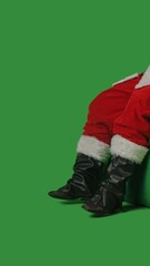 Santa Claus with a white curly beard, wearing a red suit and sunglasses, wiggles his feet while sitting on a green mockup against a green background.