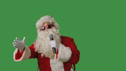 Santa Claus, with a white curly beard, wearing a red suit and sunglasses, sings songs into a microphone and dances cheerfully against a green background. Ideal for Christmas themed compositions.