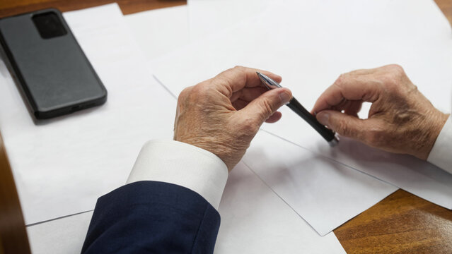 Senior businessman’s hands in a suit hold a pen over blank documents on a wooden desk; smartphone nearby, moment before signing a contract or resolution. Photo