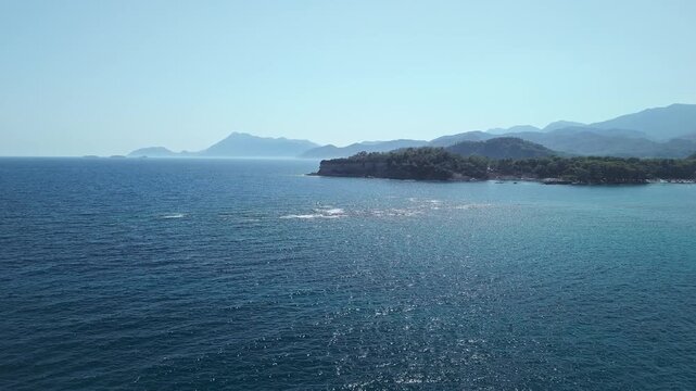 Drone view of the deep blue Mediterranean Sea with forested coastline and distant mountains near Phaselis, Antalya, Turkey. Clear summer sky and sparkling water.
