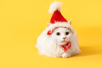 Cute white cat in funny Santa hat lying on yellow background