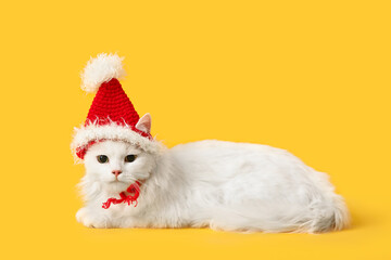 Obraz premium Cute white cat in funny Santa hat lying on yellow background
